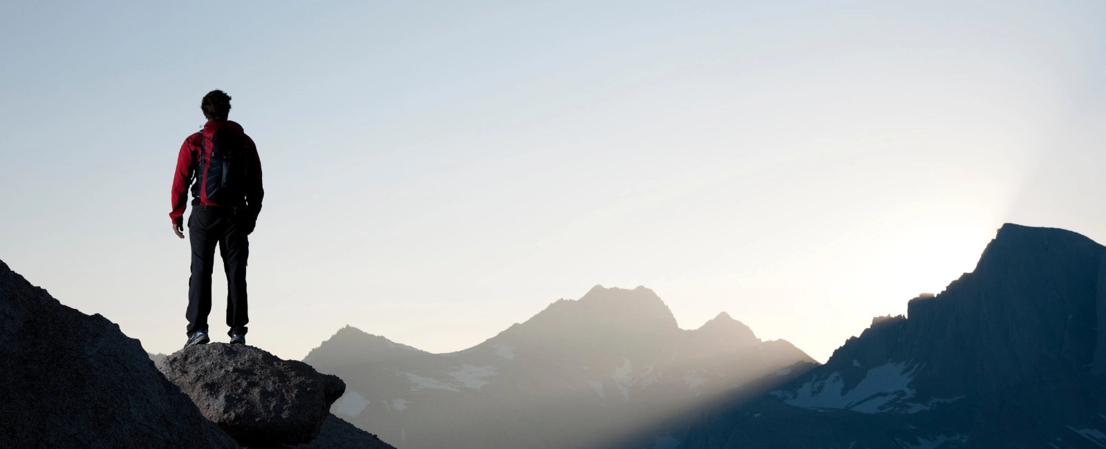 Mann mit Rucksack steht auf einem Felsen vor Sonnenaufgang in den Bergen – Symbol für Energie, Stärke und Zellgesundheit durch NMN-Supplements von Minerva-Vita.